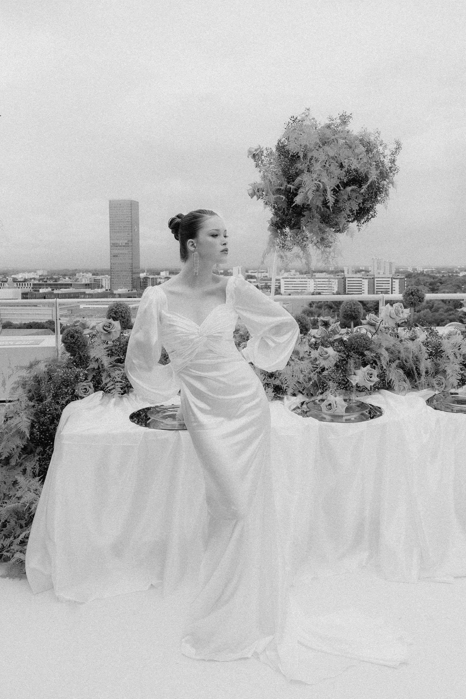 Braut in schlichtem Hochzeitskleid steht vor einem gedeckten Tisch mit Hochzeitsdekoration Tischdekoration vor der Skyline von München