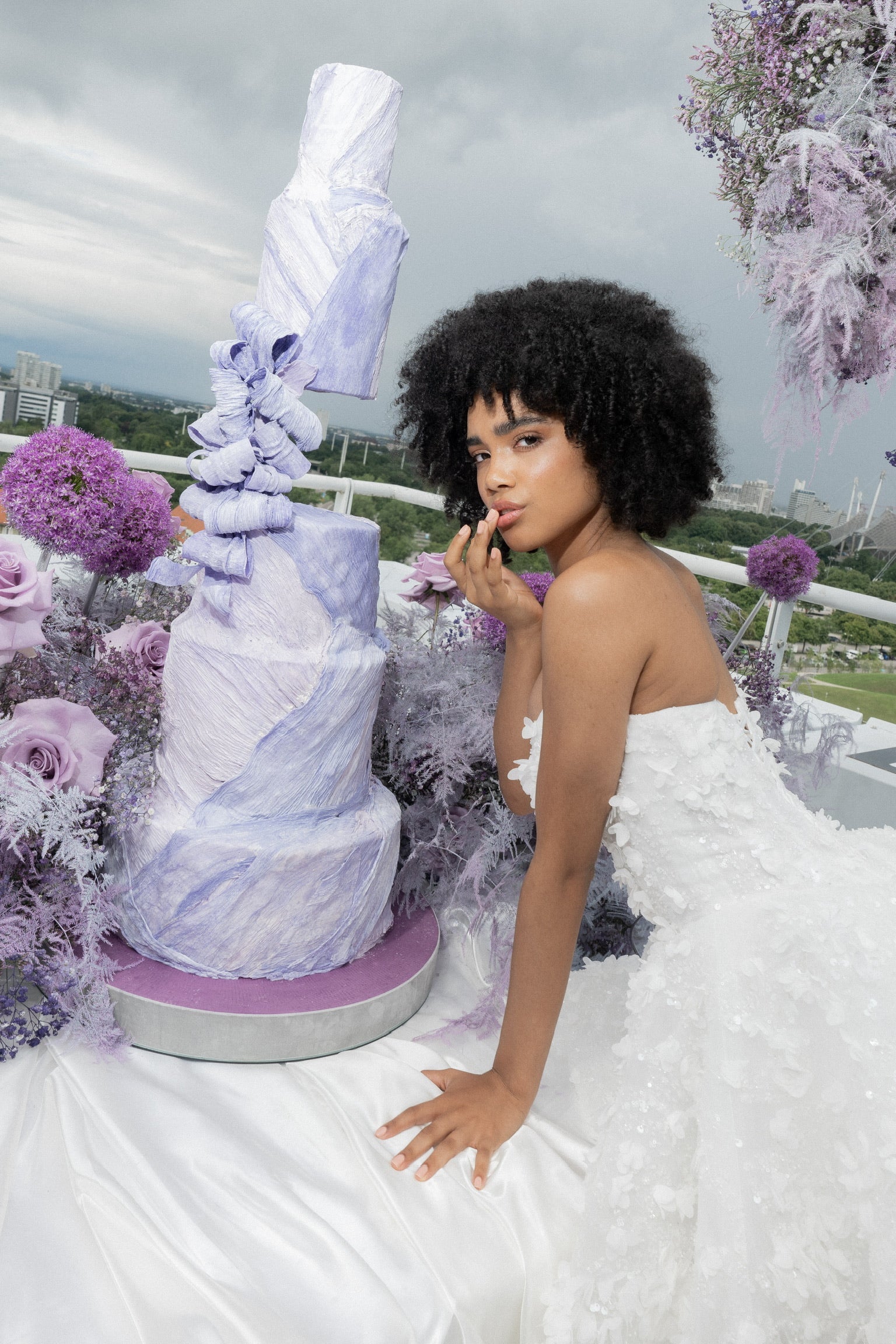 Verspielte Braut neben XXL-Hochzeitstorte mit Blumendekoration in lila-violett. Für Brautpaare mit extravaganten Vorstellungen.